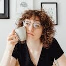 A thoughtful woman with curly hair and glasses holding a coffee mug in a modern room.