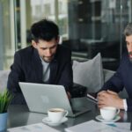 Two businessmen in a modern cafe collaborating over laptops and coffee.