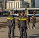 Three Dutch police officers in uniform stand in a bustling city street near public transport with people passing by.